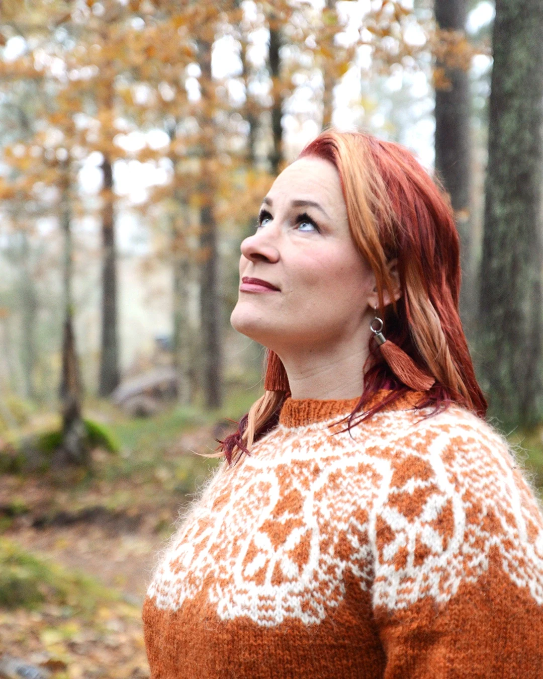 Saara katsoo ylös syksyisessä metsässä oranssissa villaneuleessa.