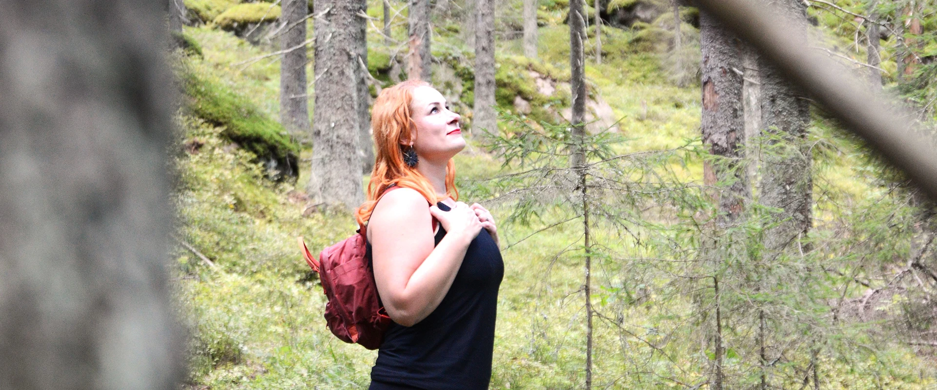 Saara seisoo kesäisessä metsässä reppu olalla ja katselee ylös puiden latvoihin.